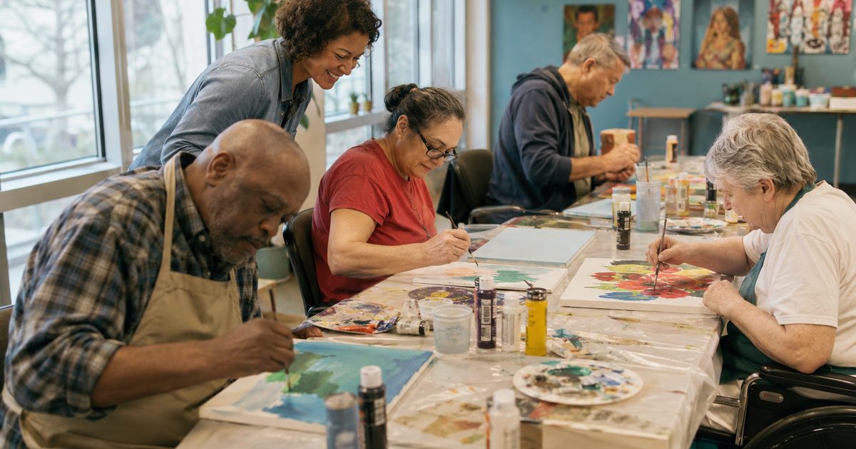 Adults painting at a long table in our art room while a staff member looks on